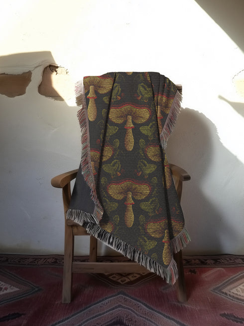 A woven cotton throw featuring an intricate mushroom and foliage pattern in deep charcoal and golden hues, framed by soft fringe.